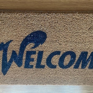 May include: A rectangular tan coir doormat featuring the word WELCOME printed in a dark blue, stylized font. The letter W is elongated with a sweeping, wave-like flourish extending to the left, while the remaining letters are in a bold, sans-serif typeface.