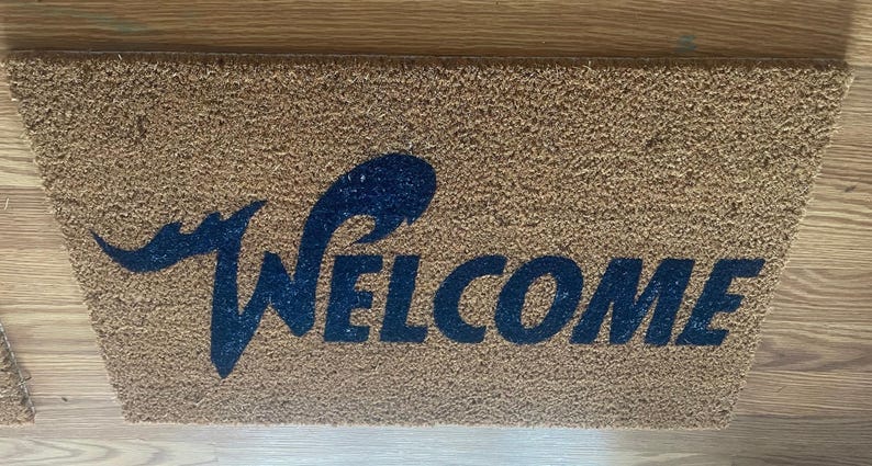 May include: A rectangular tan coir doormat featuring the word WELCOME printed in a dark blue, stylized font. The letter W is elongated with a sweeping, wave-like flourish extending to the left, while the remaining letters are in a bold, sans-serif typeface.