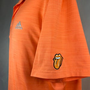 May include: Orange short-sleeved polo shirt with a silver Adidas logo on the left chest and a red, white, and black tongue logo on the right sleeve.