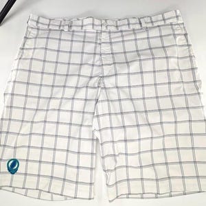 Puede incluir: Pantalones cortos blancos con un patrón de cuadros azules. Los pantalones cortos tienen cierre de botón y bolsillos. Un emblema azul y verde está bordado en la pierna izquierda.