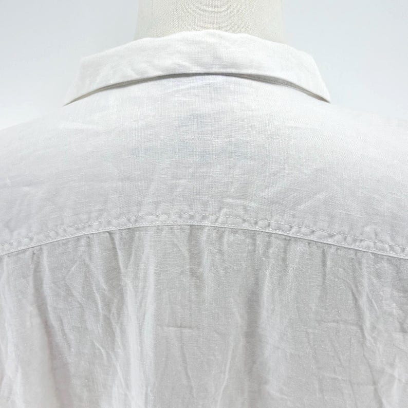 May include: Close-up of the back of a white linen shirt. The collar is visible at the top, with a yoke seam across the shoulders. The fabric has a slightly textured appearance.