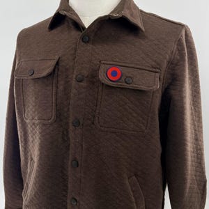 May include: Brown quilted jacket with two chest pockets and a button-down front. The jacket features a small patch with a red and blue circular design on the left chest pocket. The jacket is displayed on a white mannequin.