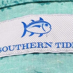 May include: A light turquoise button-down shirt with a dark teal leaf pattern. The white interior neck label features a blue fish logo, the text SOUTHERN TIDE, and a smaller tag indicating size L and that the garment was made in Indonesia.