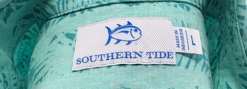 May include: A light turquoise button-down shirt with a dark teal leaf pattern. The white interior neck label features a blue fish logo, the text SOUTHERN TIDE, and a smaller tag indicating size L and that the garment was made in Indonesia.