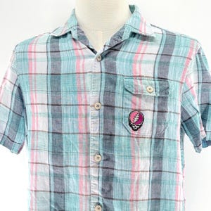 May include: Short-sleeved plaid shirt in shades of blue, pink, and white. Features a chest pocket with a colorful skull and lightning bolt patch. The shirt has a button-down front and a classic collar.