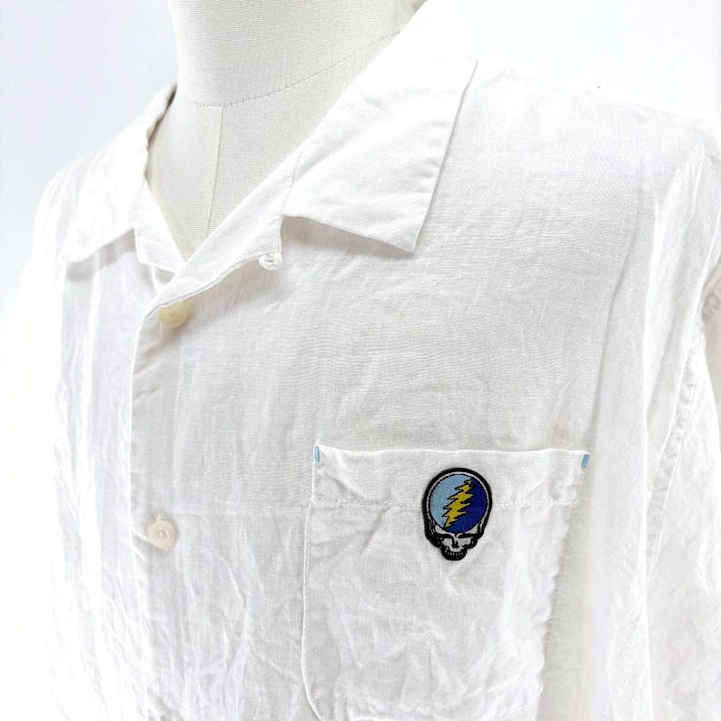 May include: White button-down shirt with a chest pocket featuring a colorful skull and lightning bolt patch. The shirt has a classic collar and button closure. The fabric appears to be lightweight.