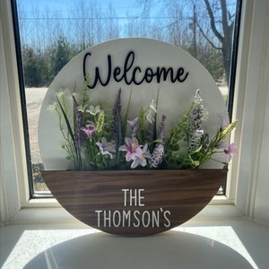 以下が含まれることがあります： 白と茶色の丸い木製看板に、黒い文字で「Welcome」と書かれており、花が飾られています。看板の下半分は茶色の木で、「The Thomson's」という文字が白い文字で書かれています。