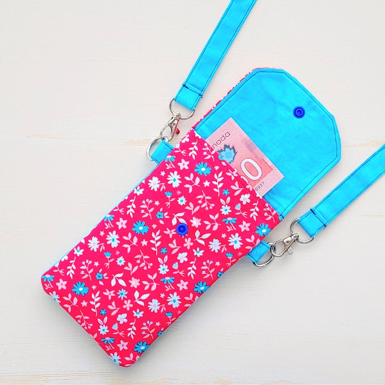 Op de afbeelding: Een roze en blauwe crossbody telefoonportemonnee met een bloemenprint en een Canadees biljet van $ 10 erin. De portemonnee heeft een blauwe flap met een drukknopsluiting.