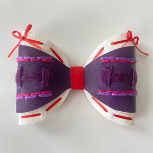 May include: A handmade bow tie with a purple leather center, white edges, and a red leather band. The purple section features embroidered details in pink and purple thread, with red stitching along the edges and red ribbon ties.