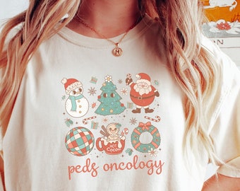Puede incluir: Camiseta color crema con un gráfico navideño colorido que incluye un muñeco de nieve, un árbol de Navidad y Papá Noel. El texto "peds oncology" está impreso debajo del diseño festivo.