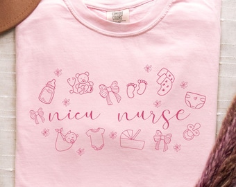 May include: Pink t-shirt with the text "NICU Nurse" in pink lettering. The shirt features a variety of small pink illustrations of baby items, including a bottle, a teddy bear, a pacifier, a diaper, and a baby onesie.