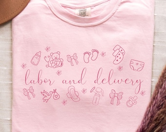 May include: Pink t-shirt with the text "labor and delivery" in pink script. The shirt is decorated with small pink illustrations of baby items, including a bottle, a teddy bear, a pacifier, a diaper, and a pair of baby shoes.