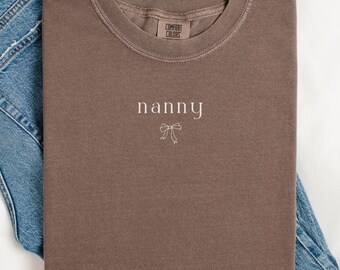May include: A brown t-shirt with the word "nanny" embroidered on the front. The shirt is folded and laid on top of a pair of blue jeans.