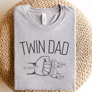May include: Gray t-shirt with the text "Twin Dad" and a graphic of two hands holding a smaller hand with the names "Sophie" and "Caleb" and the text "Est. 2024".
