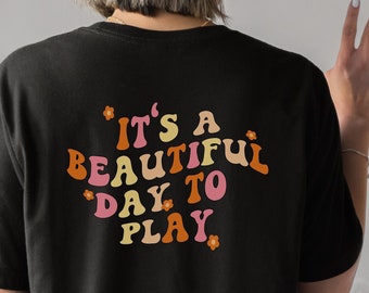 May include: Black t-shirt with the text "It's a beautiful day to play" in a retro font with pink, orange, and brown colors and small flower accents.