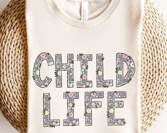May include: A cream-colored t-shirt with the words "CHILD LIFE" printed in black outlined letters. The letters are filled with a floral pattern in shades of pink, yellow, and gray.