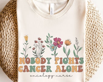May include: A white t-shirt with a floral design and the text "Nobody Fights Cancer Alone" in a retro font. The text "Oncology Nurse" is written below the main text.