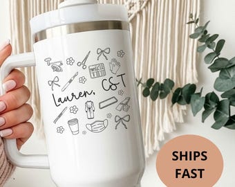 May include: White insulated tumbler with a handle, featuring black line art of scissors, flowers, and other small icons. The name "Lauren, C&T" is written in script. The tumbler has a white lid and a silver rim.