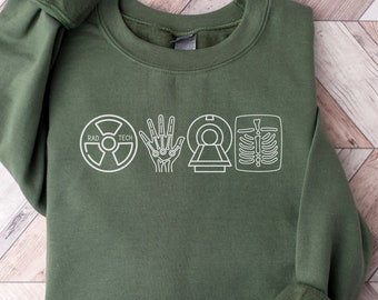 May include: A dark green sweatshirt with a white graphic design of medical imaging symbols, including a radiation symbol, a hand, a CT scan, and an X-ray. The text "Rad Tech" is printed above the symbols.