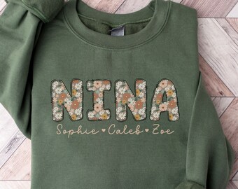 May include: A green sweatshirt with the name "NINA" in large, floral letters. The letters are outlined in black and filled with a floral pattern. Below the name, it says "Sophie Caleb Zoe" in a smaller, cursive font.