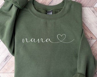 Puede incluir: Una sudadera verde oscuro con la palabra "nana" y un pequeño corazón blanco.