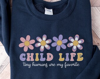 Op de afbeelding: Donkerblauwe trui met een afbeelding van roze, paarse en gele bloemen en de tekst "CHILD LIFE tiny humans are my favorite".