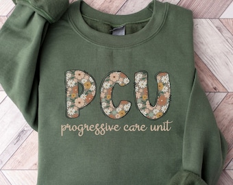 Op de afbeelding: Een groen sweatshirt met de tekst "PCU Progressive Care Unit" bedrukt op de voorkant. De letters zijn versierd met een bloemenpatroon.