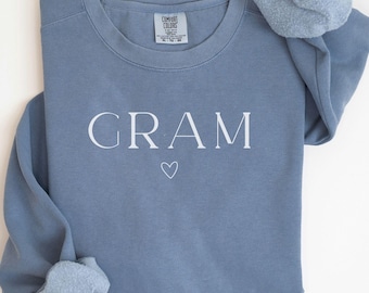 May include: A dusty blue sweatshirt with the word "GRAM" printed in white, with a small heart symbol underneath. The long-sleeved top is made of a soft material and is laid flat on a white surface.