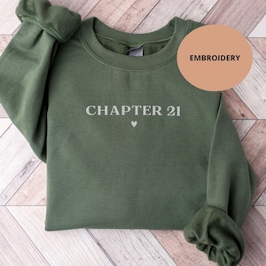 May include: An olive green sweatshirt with the text "CHAPTER 21" in white, with a small heart symbol underneath. The word "EMBROIDERY" is in a tan circle in the upper right corner. The sweatshirt is displayed on a light wood surface.