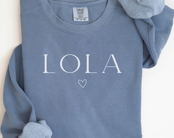 May include: A blue sweatshirt with the name "LOLA" printed in white on the front. A small white heart is below the name.