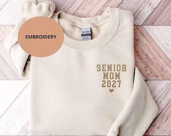 May include: A cream-colored crewneck sweatshirt featuring the embroidered text SENIOR MOM 2027 in a tan block font, positioned on the upper left chest area above a small heart icon. The garment includes a Gildan brand label at the inner neckline.