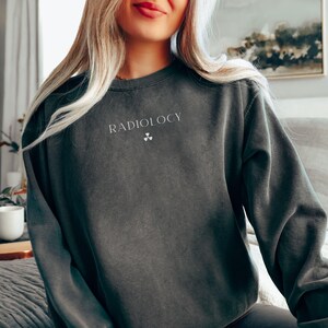 May include: Dark gray sweatshirt with the word "RADIOLOGY" printed in white, along with a radiation symbol. The sweatshirt is a solid color and has long sleeves. The image shows the top half of a person wearing the sweatshirt.