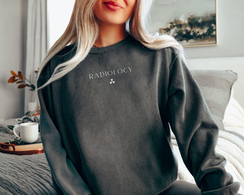 May include: Dark gray sweatshirt with the word "RADIOLOGY" printed in white, along with a radiation symbol. The sweatshirt is a solid color and has long sleeves. The image shows the top half of a person wearing the sweatshirt.