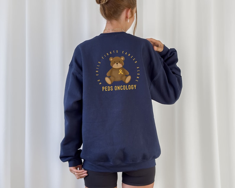 May include: Navy blue sweatshirt with a gold graphic of a teddy bear holding a gold ribbon. The text around the bear reads "No Child Fights Cancer Alone & Peds Oncology".