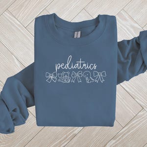 K&ouml;nnte beinhalten: Ein blaues Sweatshirt mit dem Wort "P&auml;diatrie" in wei&szlig;er Schrift. Das Wort ist umgeben von kleinen wei&szlig;en Illustrationen von Schleifen, einem Stethoskop, einem Teddyb&auml;r, einem Dinosaurier und einem Schnuller.