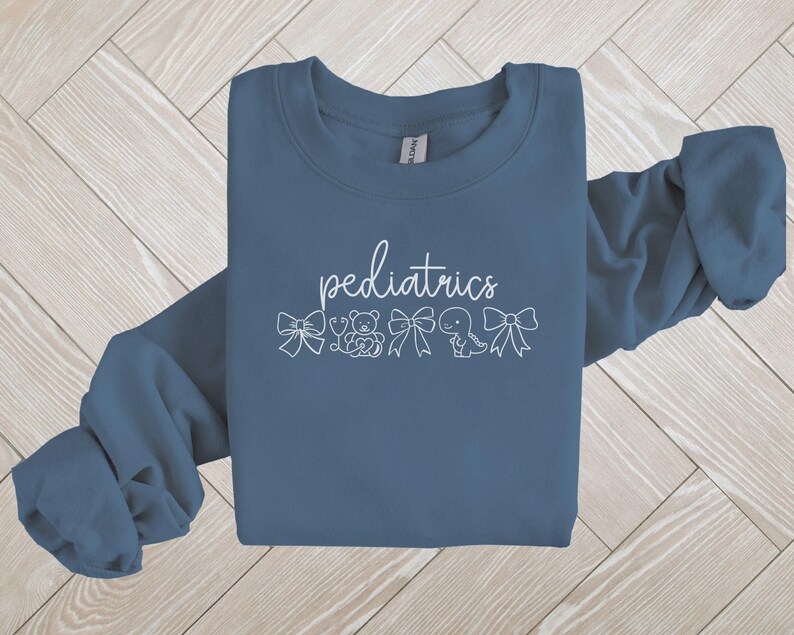K&ouml;nnte beinhalten: Ein blaues Sweatshirt mit dem Wort "P&auml;diatrie" in wei&szlig;er Schrift. Das Wort ist umgeben von kleinen wei&szlig;en Illustrationen von Schleifen, einem Stethoskop, einem Teddyb&auml;r, einem Dinosaurier und einem Schnuller.
