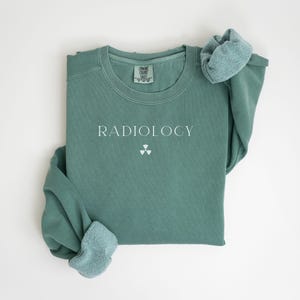 May include: A sage green sweatshirt with the word "RADIOLOGY" printed in white, along with a radiation symbol. The long-sleeved shirt has a crew neck and a soft, comfortable appearance. The sleeves are folded over.