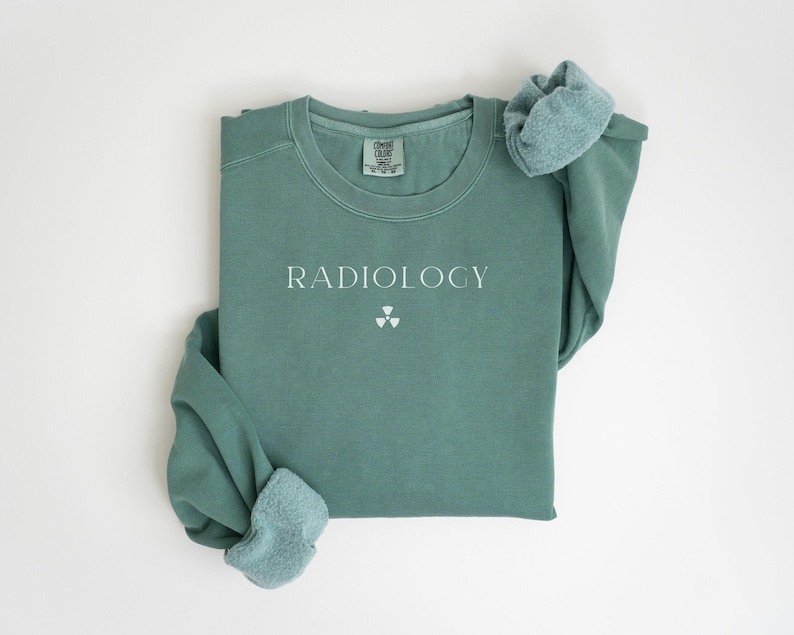 May include: A sage green sweatshirt with the word "RADIOLOGY" printed in white, along with a radiation symbol. The long-sleeved shirt has a crew neck and a soft, comfortable appearance. The sleeves are folded over.