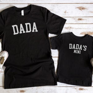 May include: Two black t-shirts with white text. The larger shirt says "DADA" in a collegiate font. The smaller shirt says "DADA'S MINI" in the same font.