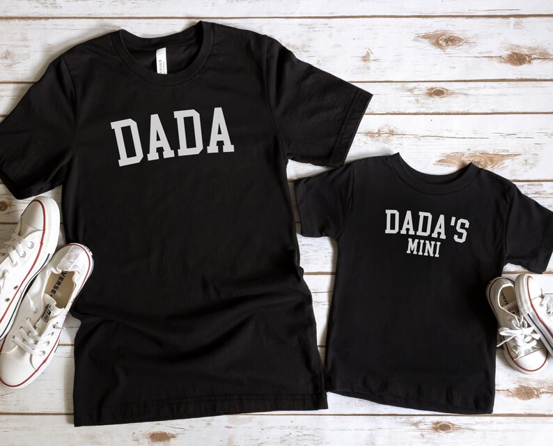 May include: Two black t-shirts with white text. The larger shirt says "DADA" in a collegiate font. The smaller shirt says "DADA'S MINI" in the same font.