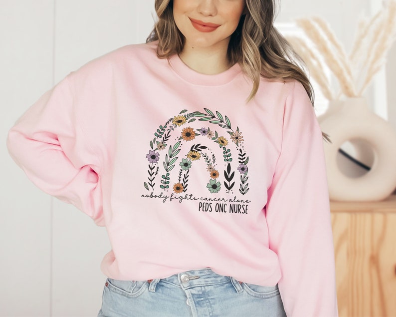 Peut inclure: Sweat-shirt rose avec un arc-en-ciel floral et le texte "Personne ne se bat contre le cancer seul. Infirmi&egrave;re en oncologie p&eacute;diatrique."