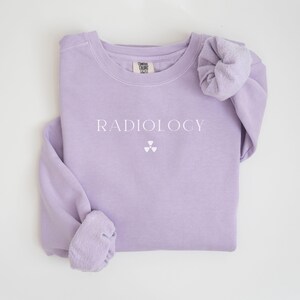 May include: A light purple sweatshirt with the word "RADIOLOGY" printed in white, along with a white radiation symbol. The long-sleeved shirt is folded neatly, showcasing the design on the front.