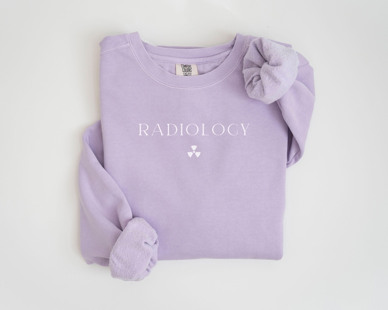 May include: A light purple sweatshirt with the word "RADIOLOGY" printed in white, along with a white radiation symbol. The long-sleeved shirt is folded neatly, showcasing the design on the front.