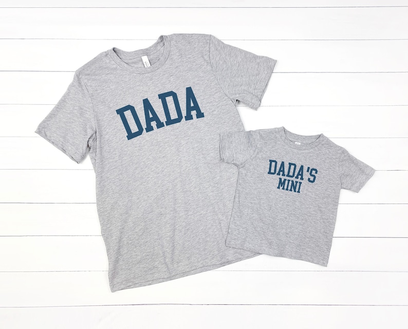 May include: Two gray t-shirts with blue text. The larger shirt says "DADA" and the smaller shirt says "DADA'S MINI".