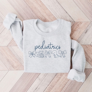 K&ouml;nnte beinhalten: Ein grauer Pullover mit dem Wort "P&auml;diatrie" in Blau bedruckt. Das Wort ist von kleinen blauen Illustrationen eines Stethoskops, eines Teddyb&auml;ren, einer Schleife, eines Dinosauriers und einer weiteren Schleife umgeben.