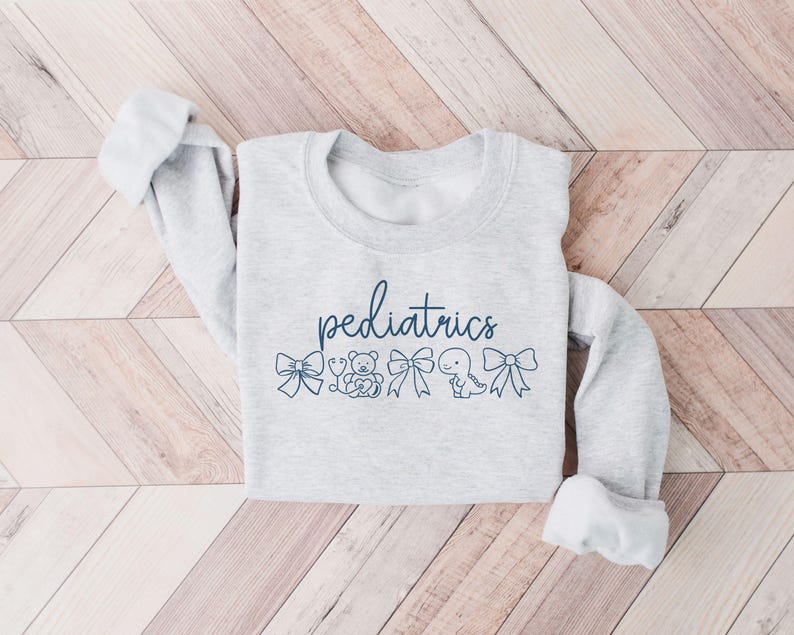 K&ouml;nnte beinhalten: Ein grauer Pullover mit dem Wort "P&auml;diatrie" in Blau bedruckt. Das Wort ist von kleinen blauen Illustrationen eines Stethoskops, eines Teddyb&auml;ren, einer Schleife, eines Dinosauriers und einer weiteren Schleife umgeben.