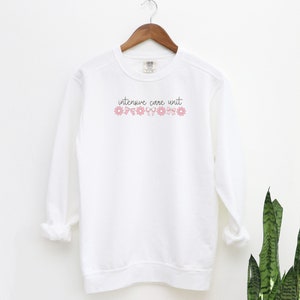 May include: White sweatshirt with the text "intensive care unit" in black script with pink flowers and bows above the text.