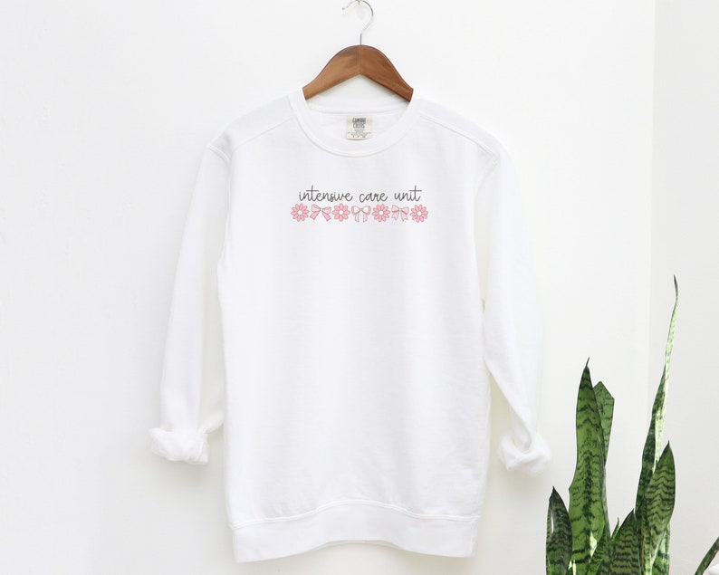 May include: White sweatshirt with the text "intensive care unit" in black script with pink flowers and bows above the text.