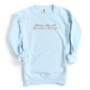 May include: Light blue sweatshirt with the text "intensive care unit" in black lettering and pink flowers and bows.