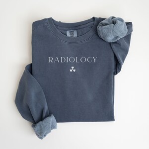 May include: A slate blue sweatshirt with the word "RADIOLOGY" printed in white, along with a radiation symbol. The sleeves are cuffed, and the garment is laid flat on a white surface. The sweatshirt is a Comfort Colors brand.